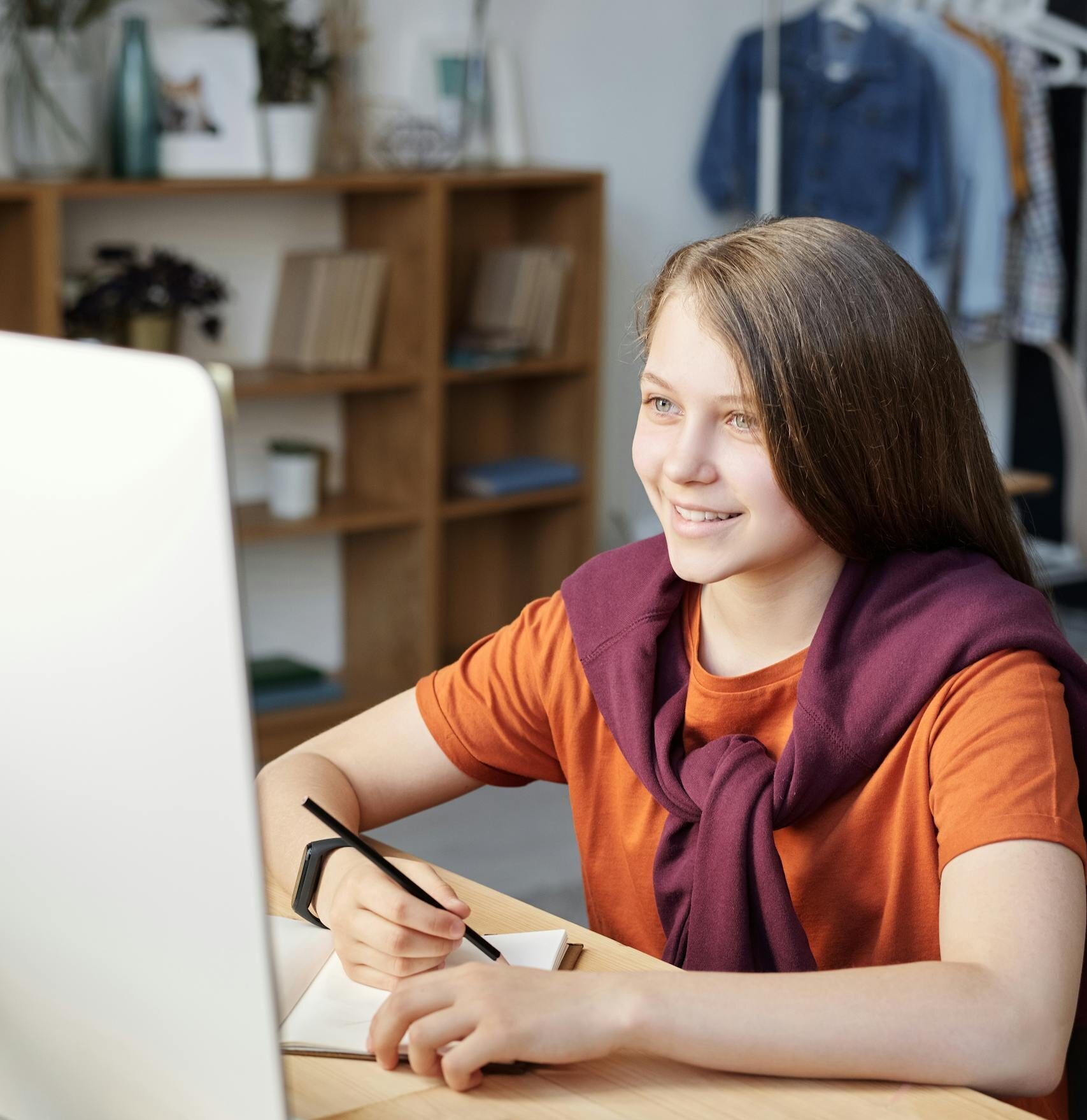 Smiling teen girl studying online at her home desk with a computer and notepad.